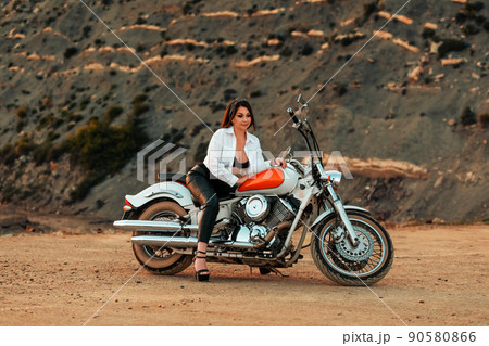 A beautiful adult woman with a high heels, sexy posing sitting on motorcycle. Rock on the background. The concept of Motorcyclist Day A beautiful adult woman with a high heels, sexy posing sitting on motorcycle. Rock on the background. The concept of Motorcyclist Day 90580866
