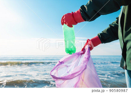A volunteer wearing rubber gloves puts a dirty plastic bottle in a garbage bag. In the background is the ocean and the horizon line. Hands close-up. Earth Day concept 90580987