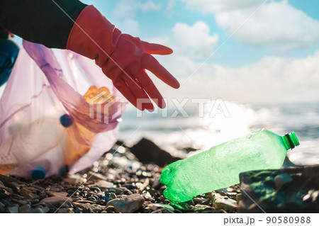 Earth Day. A person in protective rubber gloves reaches for a dirty plastic green bottle lying on the ocean shore. Hand close-up. Copy space. Concept of save the Planet Earth Day. A person in protective rubber gloves reaches for a dirty plastic green bottle lying on the ocean shore. Hand close-up. Copy space. Concept of save the Planet 90580988