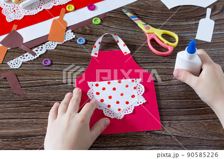 A child is gluing paper details of a gift from a paper apron postcard on mother's day. Hand-made. The project of children's creativity, needlework, crafts for children. 90585226