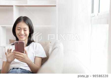Asian woman sitting with smart phone on sofa in living room, Looking at tablet screen, Relaxing at home, Working through the Internet communication system, Happy working lifestyle. 90586220