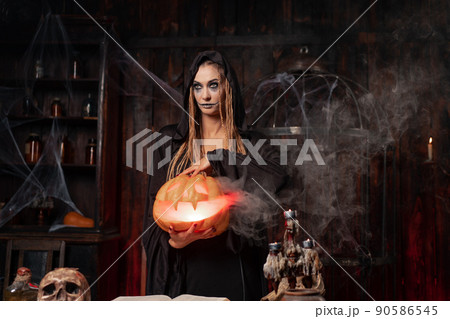 Halloween concept. Black witch holding Halloween pumpkin with carved smily face in hand standing dark room Halloween concept. Black witch holding Halloween pumpkin with carved smily face in hand standing dark room 90586545