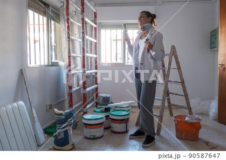 Young woman painting interior wall with white paint. paint cans and painter ladder. Young woman painting interior wall with white paint. paint cans and painter ladder. 90587647