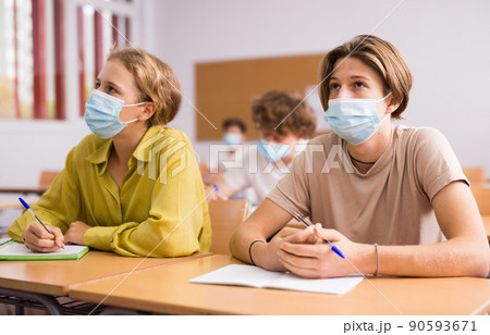 Teenager in protective mask studying in classroom, listening to lecturer and writing in notebook 90593671