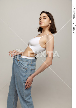 Portrait of young slim wom,an posing in white top and oversized jeans isolated oevr grey studio background. Weight-loss 90606736