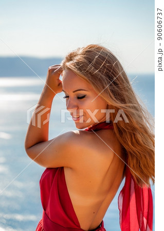 Smiling young woman in a red dress looks at the camera. A beautiful tanned girl enjoys her summer holidays at the sea. Portrait of a stylish carefree woman laughing at the ocean. Smiling young woman in a red dress looks at the camera. A beautiful tanned girl enjoys her summer holidays at the sea. Portrait of a stylish carefree woman laughing at the ocean. 90606737