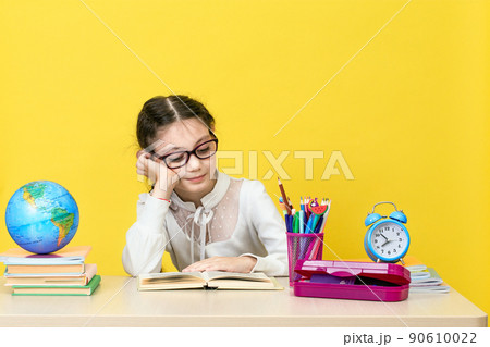 The schoolgirl sits at the desk and thinks about the decision of the task on yellow background. 90610022