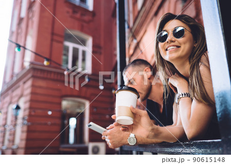 Joyful woman in sunglasses posing near house holding disposable takeaway coffee Joyful woman in sunglasses posing near house holding disposable takeaway coffee 90615148