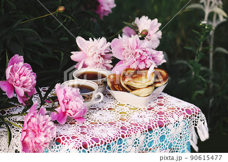 Tea in country style in summer garden. Two cups of black tea and pancakes on handmade crocheted vintage lacy tablecloth and blooming peony flowers in sunlight. Tea in country style in summer garden. Two cups of black tea and pancakes on handmade crocheted vintage lacy tablecloth and blooming peony flowers in sunlight. 90615477