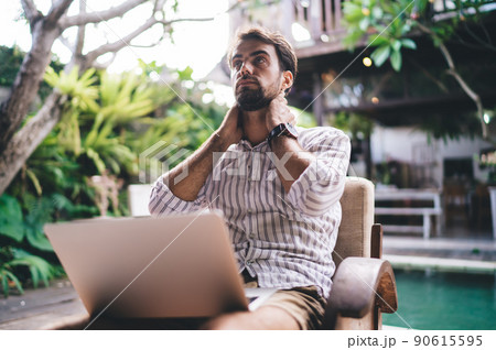 Young serious businessman thinking while touching neck and using laptop 90615595