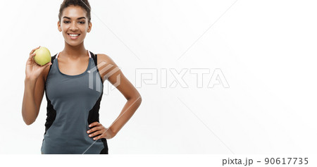 Healthy and Fitness concept - Beautiful American African lady in grey fitness clothes holding green apple. Isolated on white background. Healthy and Fitness concept - Beautiful American African lady in grey fitness clothes holding green apple. Isolated on white background. 90617735