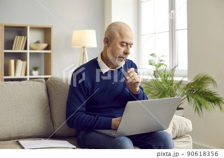 Senior man sitting on couch at home, working on his laptop computer and thinking Senior man sitting on couch at home, working on his laptop computer and thinking 90618356