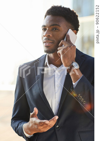 Content modern young Afro-American manager with beard standing outdoors and gesturing hand while communicating on phone Content modern young Afro-American manager with beard standing outdoors and gesturing hand while communicating on phone 90622152