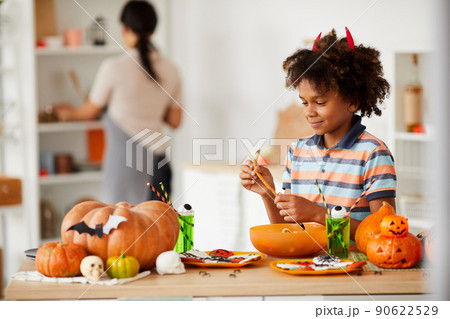 Smiling cute black boy with devils horns standing at counter with various Halloween candies and eating marmalade worm Smiling cute black boy with devils horns standing at counter with various Halloween candies and eating marmalade worm 90622529