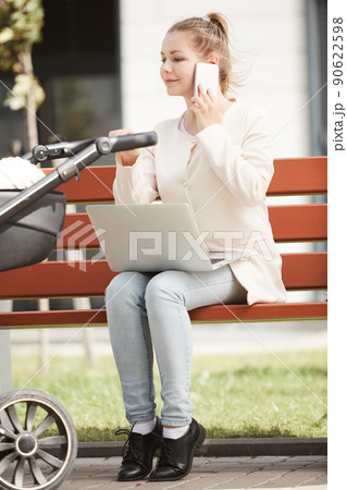 Young mother in white coat working outside on her smartphone and laptop while baby sleeping in stroller Young mother in white coat working outside on her smartphone and laptop while baby sleeping in stroller 90622598