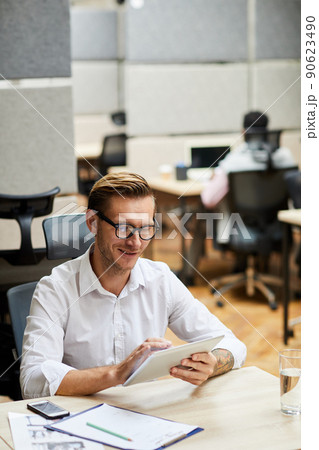 Positive handsome manager with fashion hairstyle sitting at desk and viewing sales data on tablet Positive handsome manager with fashion hairstyle sitting at desk and viewing sales data on tablet 90623490