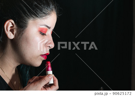 beautiful latina woman using a brush to paint her lips red, girl with her eyes painted with red shadow. with space for advertising text. black background. beauty concept. 90625172