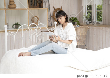 ベッドに座ってタブレットを見る若い女性 90626015