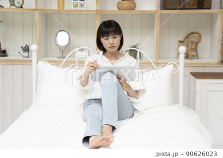ベッドに座ってタブレットを見る若い女性 90626023