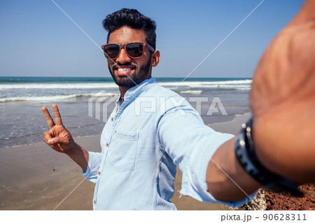 Young african american man in blue shirt and jeans making a selfie portrait on the front camera of a smartphone on the ocean beach. 90628311
