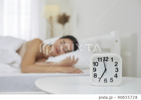 Alarm clock set for 7 AM on bedside table, with happy woman sleeping on bed in background 90655728