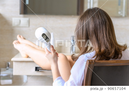 Back view of woman in a white nightgown blow dry hair by hair dryer sitting on armchair and looking in front of the mirror in the bathroom 90657104