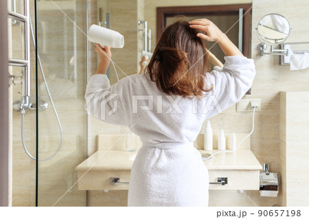 Back view of woman in a white robe blow dry hair by hair dryer standing and looking in front of the mirror in the bathroom 90657198