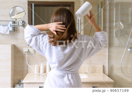 Back view of woman in a white robe blow dry hair by hair dryer standing and looking in front of the mirror in the bathroom 90657199