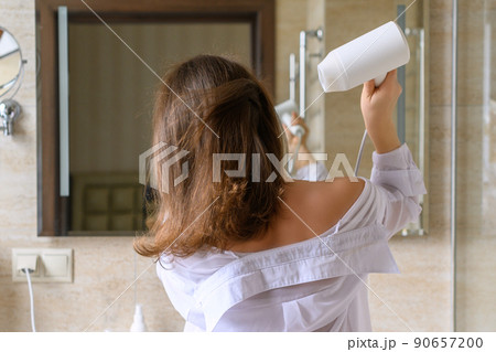 Back view of woman in a white nightgown blow dry hair by hair dryer standing and looking in front of the mirror in the bathroom 90657200