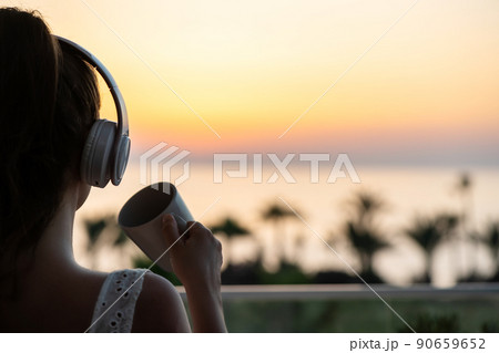 Rear view of woman with headphones and cup of tea looking on palm trees sea beach at sunset. Female relaxing listenning music. Rear view of woman with headphones and cup of tea looking on palm trees sea beach at sunset. Female relaxing listenning music. 90659652