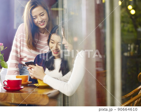 three young asian women looking at mobile phone in coffee shop three young asian women looking at mobile phone in coffee shop 90664610