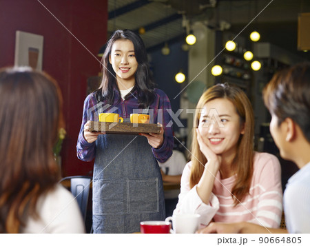 young smiling asian waitress serving customers in coffee shop, focus on the background girl young smiling asian waitress serving customers in coffee shop, focus on the background girl 90664805