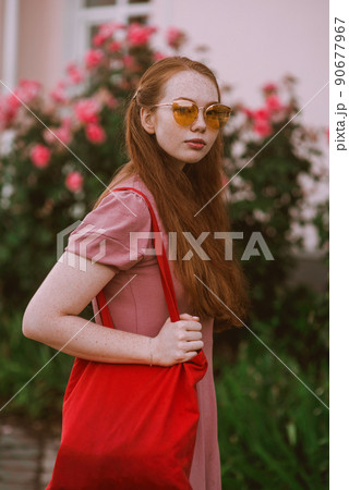 Young beautiful red-haired long-haired woman in a summer pink dress walks through the streets of a summer city. Young beautiful red-haired long-haired woman in a summer pink dress walks through the streets of a summer city. 90677967