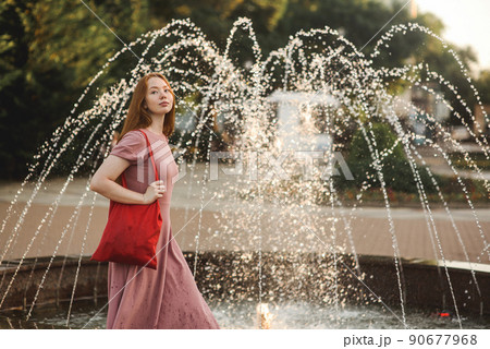 Young beautiful red-haired long-haired woman in a summer pink dress walks through the streets of a summer city. Young beautiful red-haired long-haired woman in a summer pink dress walks through the streets of a summer city. 90677968