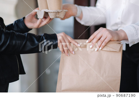Customer in face mask stand outdoors and take package with coffee and food from cashier in mask at cafe. Take away food. Close up of hands. Customer in face mask stand outdoors and take package with coffee and food from cashier in mask at cafe. Take away food. Close up of hands. 90687190