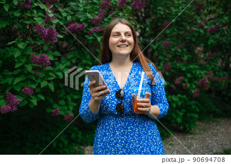 Young beautiful woman in bright blue dress holding plastic cold tea cup and looking at smartphone while walking at city park. Businesswoman drinking cold cocktail and smiling. Young beautiful woman in bright blue dress holding plastic cold tea cup and looking at smartphone while walking at city park. Businesswoman drinking cold cocktail and smiling. 90694708