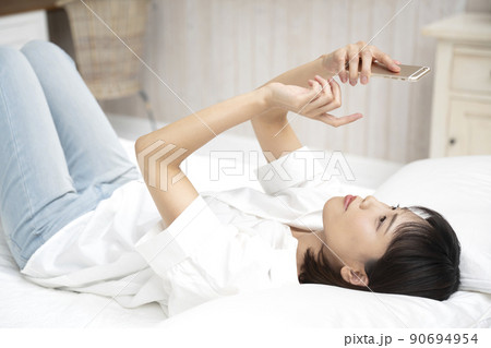 ベッドに仰向けで寝てスマホの操作をする若い女性 90694954