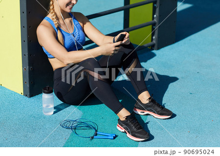 Positive girl in black leggings sitting at sports ground and monitoring her training on phone while analyzing her progress Positive girl in black leggings sitting at sports ground and monitoring her training on phone while analyzing her progress 90695024