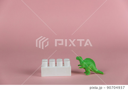 A dinosaur miniature and a white block of children's constructor against a pink background. Small green miniature of a predatory dinosaur. Close-up. Selective focus. A dinosaur miniature and a white block of children's constructor against a pink background. Small green miniature of a predatory dinosaur. Close-up. Selective focus. 90704937