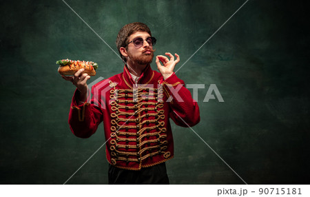 Young man in suit as royal hussar tasting big hot-dog isolated on dark green background. Retro style, comparison of eras concept. Young man in suit as royal hussar tasting big hot-dog isolated on dark green background. Retro style, comparison of eras concept. 90715181