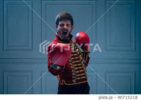 Emotional young man like medieval hussar wearing boxing gloves isolated on dark blue background. Retro style, hobby, comparison of eras concept. 90715218