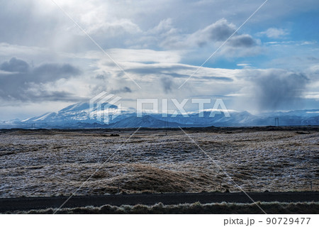 View of road by grassy landscape and snow covered mountains against cloudy sky 90729477