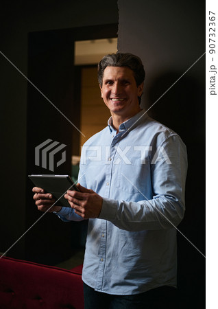 Handsome successful mature Caucasian man holds digital gadget and smiles looking at camera. Interior designer, entrepreneur in a furniture design showroom. 90732367