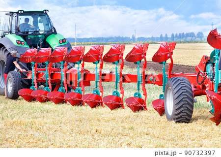 Red plow as a trailer on a tractor on the background of an agricultural field. 90732397