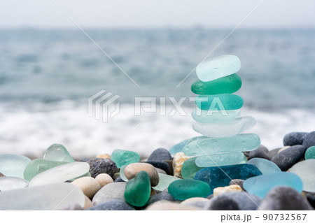 Natural polish textured sea glass and stones on the seashore. Azure clear sea water with waves. Green, blue shiny glass with multi-colored sea pebbles close-up. Beach summer background.  90732567