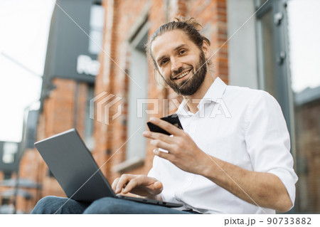 Stylish bearded man doing online purchases while sitting outside near office building. Stylish bearded man doing online purchases while sitting outside near office building. 90733882