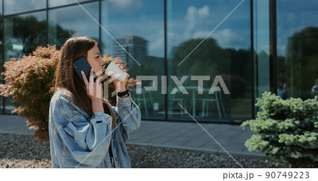 Beautiful woman wearing in a denim jacket and using smartphone while walking in the city. Woman talking on phone with friends rejoicing smiling laughing and drinks coffee. Beautiful woman wearing in a denim jacket and using smartphone while walking in the city. Woman talking on phone with friends rejoicing smiling laughing and drinks coffee. 90749223