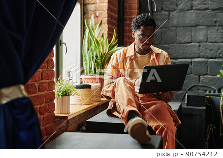 Young black man in casualwear sitting by window and neworking on laptop 90754212