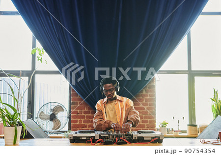 Young man in headphones and casualwear standing by table with dj set Young man in headphones and casualwear standing by table with dj set 90754354