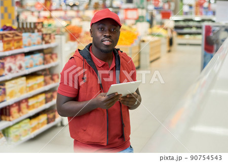 African American guy in red uniform and cap using digital tablet in supermarket 90754543